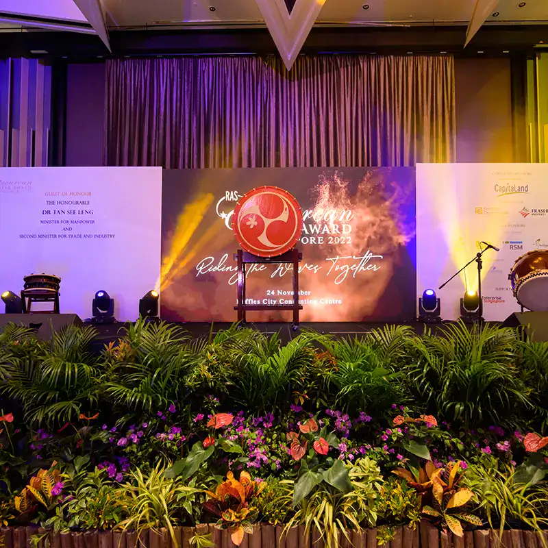 Elegant stage setup for awards event with vibrant lighting and lush floral arrangements by The Project Crew